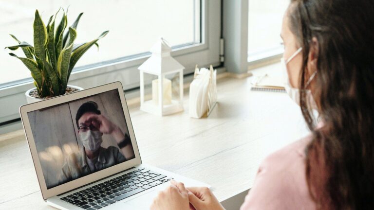 Imagem de uma mulher com máscara usando o computador para fazer uma ligação em vídeo com outro colega de trabalho, representando o post sobre saúde mental na pandemia