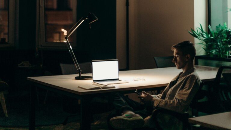 Imagem de um homem trabalhando à noite, com um notebook e um celular na mão. A imagem representa nosso conteúdo sobre abandono de emprego