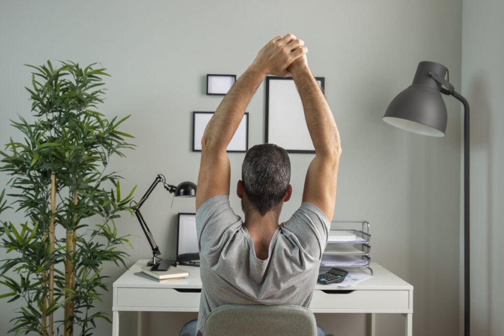 Um homem está trabalhando em casa, visto de costas. Ele está sentado à sua escrivaninha, com uma postura focada enquanto se dedica a suas tarefas.